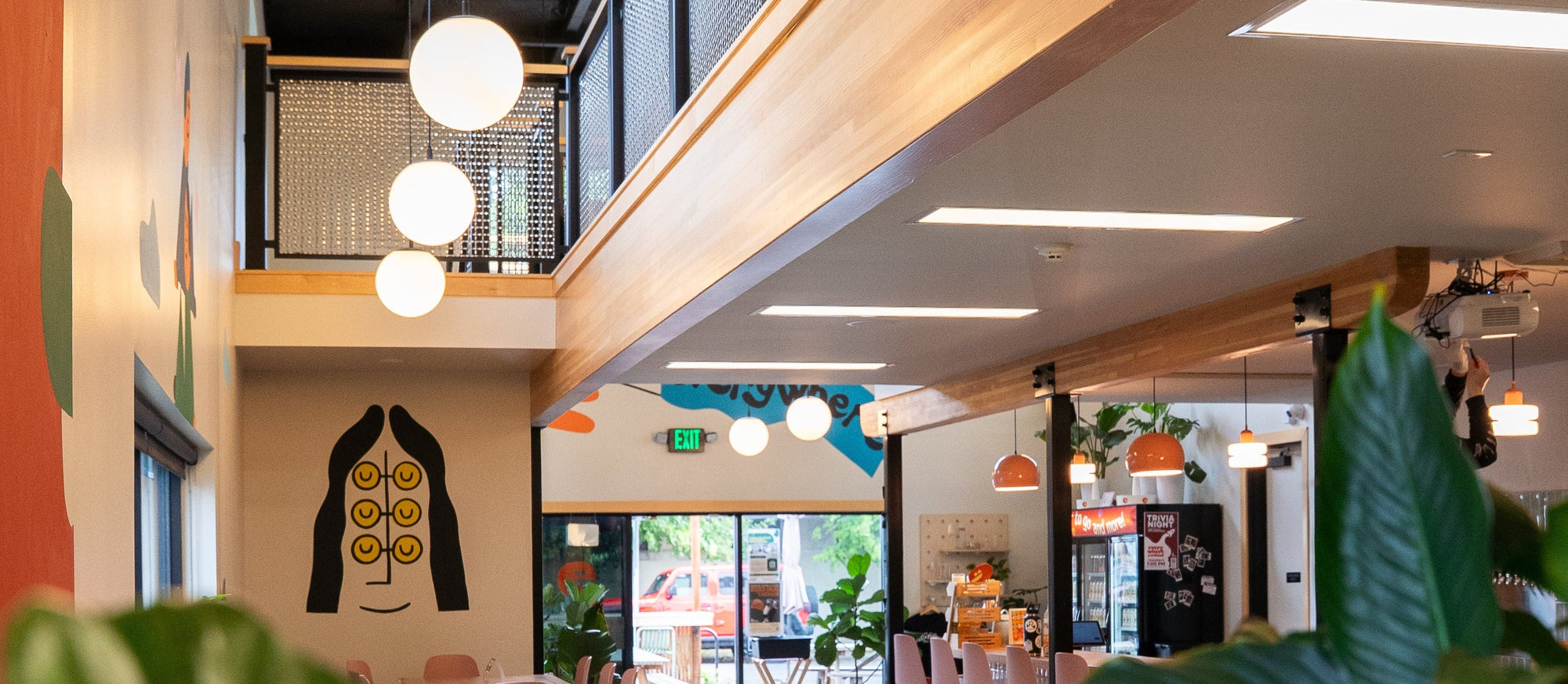 A modern, two-story office or cafe interior with a wooden ceiling, black mesh upper railing, hanging white globe lights, and numerous green houseplants in the foreground. The space features colorful walls, a black office chair, and a glass door in the distance.