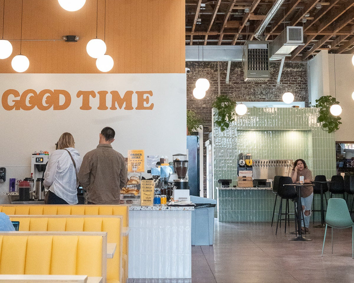 A bustling cafe interior with a counter where two people are ordering, yellow booth seating in the foreground, exposed wooden ceiling beams, hanging globe lights, and green trailing plants throughout the space.